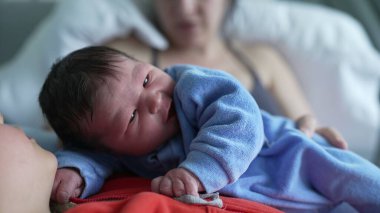 Mavi tulumlu yeni doğmuş bir bebeğin hassas anı. Arka planda anne ile kardeşinin göğsünde dinleniyor. Huzurlu ve samimi bir ortamda sevgi ve aile bağını yakalıyor.