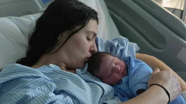 stock image Mother kissing newborn baby infant right after birth, initial day of infant being held and cared by mom at hospital clinic cradling son in arms