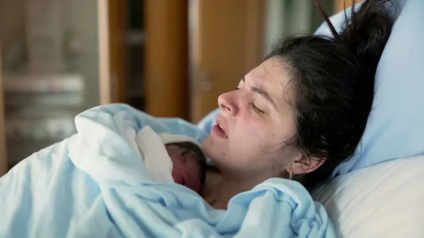 stock image New mother in discomfort after post-partum trauma at hospital clinic holding newborn on chest. woman in pain after childbirth, real life authentic emotion of human struggle