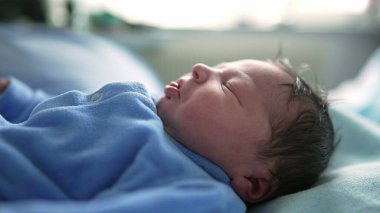 Yeni doğan bebek huzur içinde uyuyor mavi tulum giyiyor, bebeğin yüzüne odaklanıyor. Huzurlu ifade, yeni doğan bir bebeğin çocukluğunda huzurlu uykusunun masumiyetini ve huzurunu vurgular.