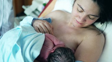 Yenidoğan 'ın İlk Beslenmesi- Hemşire bebeğini battaniyeyle örterken, anne de hastane kliniğinde cilt cilde teması vurguluyor.