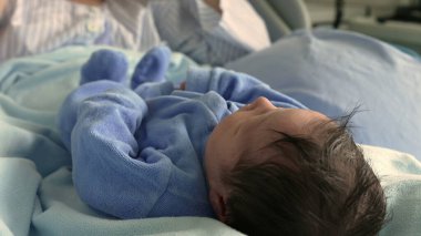 Yeni doğmuş bir bebeğin mavi bir tulumun içinde huzurlu bir şekilde uyuması. Sakin ifade ve nazik yüz hatları, yeni doğan bir bebeğin ilk günlerindeki sükunet ve masumiyetini vurgular. Saf bir sükunet anını yakalar.