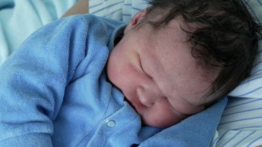 Yeni doğmuş bebek mavi bir tulumla huzur içinde uyuyor, sarılıp sarmalanmış ve rahat. Bebeklerin yüzündeki dingin ifade bu hassas anda çocukluğun masumiyetini ve sakinliğini vurgular.
