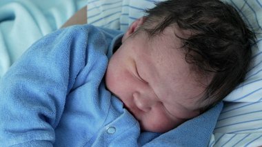 Yeni doğmuş bebek mavi bir tulumla huzur içinde uyuyor, sarılıp sarmalanmış ve rahat. Bebeklerin yüzündeki dingin ifade bu hassas anda çocukluğun masumiyetini ve sakinliğini vurgular.