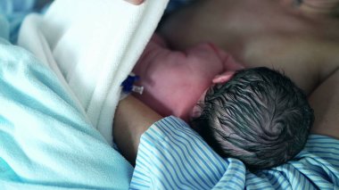 Yenidoğan 'ın İlk Beslenmesi- Hemşire bebeğini battaniyeyle örterken, anne de hastane kliniğinde cilt cilde teması vurguluyor.