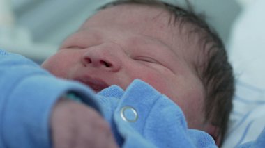 Mavi tulumlu yeni doğmuş bir bebeğin gözleri kapalı bir şekilde huzurlu bir şekilde uyuması. Yumuşak cilt ve gevşek ifade, çocukluk döneminin huzurlu ve hassas anlarını vurgular.