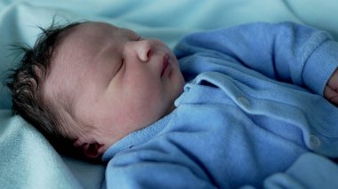 Yumuşak, koyu renk saçlı yeni doğmuş bebek, huzur içinde mavi bir tulumla uyuyor, bir hastane yatağında, yumuşak ışık ve mavi tonlarda, çocukluk döneminin masumiyetini ve sükunetini vurguluyor.