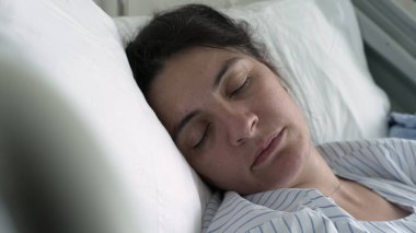 Anne bir hastane yatağında huzur içinde dinleniyor, gözleri kapalı, çizgili bir hastane önlüğü giyiyor, doğum sonrası huzurlu ve hassas anlar geçiriyor, anne dayanıklılığını ve iyileşmesini vurguluyor.