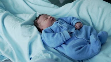 Yumuşak, koyu renk saçlı yeni doğmuş bebek, huzur içinde mavi bir tulumla uyuyor, bir hastane yatağında, yumuşak ışık ve mavi tonlarda, çocukluk döneminin masumiyetini ve sükunetini vurguluyor.