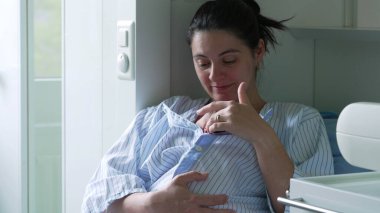 Anne hastane odasında bebeğini göğsüne yakın tutarken gülümsüyor. Mavi ve beyaz çizgili tişörte sarılmış bebek annesinin sıcaklığından ve güvenliğinden zevk alır.