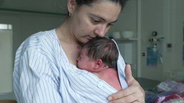 Anne yeni doğmuş bebeğini bir hastane odasında göğsüne yakın tutarak ona sıcaklık ve rahatlık sağlıyor. Çizgili bir tişörte sarılı yeni doğan anne ve çocuk arasındaki bağı gözler önüne sererek huzur içinde uyuyor.