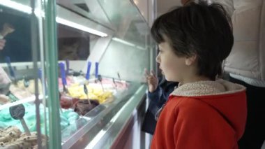 Çocuk dondurmacıda dondurma seçiyor, vitrindeki renkli tatlara bakıyor, dondurma salonu, karar verme anı, bardaktaki yansıma, tatlı seçimi