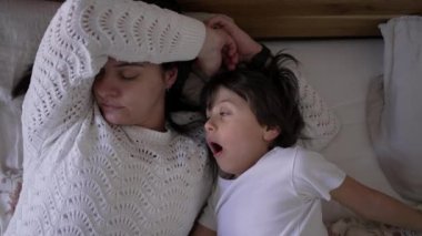 Anne ve küçük çocuk yatakta yatıyor, çocuk esniyor ve anne dinleniyor, hassas ve rahat bir aile anını yakalıyor, aile bağlarını ve günlük rutinlerini ön plana çıkaran yaşam tarzı için mükemmel.