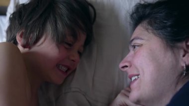 Anne ve küçük oğlu gülüyor ve yatakta neşeli bir anı paylaşıyorlar anne oğlunun neşeli yüz ifadelerine gülümsüyor aile bağlarını güçlendirmek için sabah vakti ve mutluluğu.