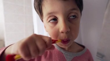 Dişlerini renkli bir diş fırçasıyla fırçalayan, çizgili pijama giyen, iyi aydınlatılmış bir banyoda ağız temizliği alışkanlıkları ve çocukluk rutinlerine odaklanan genç bir çocuk.