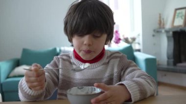Genç çocuk masada bir kase yoğurt yiyor, kaşığı düşünceli bir ifadeyle ağzına doğru kaldırıyor, ev ortamında sakin ve samimi bir anı yakalıyor.