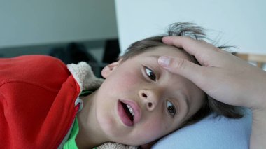 Kırmızı kapüşonlu, tuhaf bir ifadeyle uzanmış, annesinin elinden nazikçe kafa masajı alan, özen, rahatlama ve aile bağlarını resmeden genç bir çocuğun yakın çekimi.