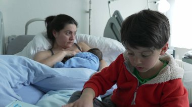 Mother breastfeeding newborn while older sibling in red jacket sits nearby on hospital bed. Mother's expression shows fatigue, and child plays by himself. Family bond in hospital