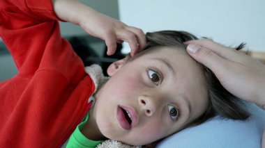 Kırmızı kapüşonlu, tuhaf bir ifadeyle uzanmış, annesinin elinden nazikçe kafa masajı alan, özen, rahatlama ve aile bağlarını resmeden genç bir çocuğun yakın çekimi.