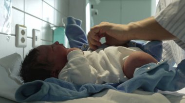 Yeni doğmuş bir bebeğin hastanede mavi tulum giymesi, doğumdan hemen sonra bebeklere sunulan şefkati ve sıcaklığı gösteriyor.