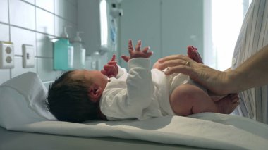 Yeni doğmuş bir bebek, ebeveynleri tarafından beyaz bir kıyafet giydiriliyor, hastane masasında uzanıyor, doğumdan hemen sonra bebeklere verilen bakımı vurguluyor.