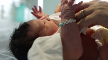 Yeni doğmuş bebek isimlendirilmiş bilezik takıyor, hastanede ebeveyn tarafından giydiriliyor, isim boncuklarıyla küçük el ve bileklere yakın çekim yapıyor, sevgi ve özeni simgeliyor.