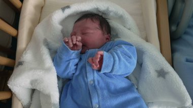 Newborn baby in blue onesie crying in crib, expression of discomfort, soft blanket, early days of life, capturing raw emotions, detailed close-up of baby's face, first moments, infancy