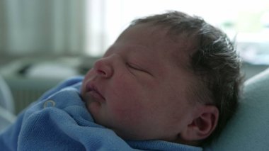 Side profile of a newborn baby in a blue onesie, peacefully asleep in a parent's arms, capturing the tender and serene moments of early parenthood and the beginning of a new life