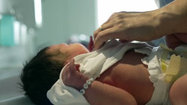 Koyu renk saçlı yeni doğmuş bebek hastanede ebeveyni tarafından nazikçe giydiriliyor. Küçük kafa ve boynuna yakın plan çekiliyor. İlk anlarda hassas bir anne-baba bakımı sergiliyor.