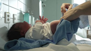 Yeni doğmuş bir bebeğin hastanede mavi tulum giymesi, doğumdan hemen sonra bebeklere sunulan şefkati ve sıcaklığı gösteriyor.