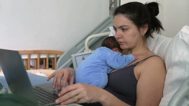 Mother in hospital bed working on laptop with newborn baby on chest, blue onesie, multitasking, new parent, early motherhood, maternal care, balancing work and baby, bonding time