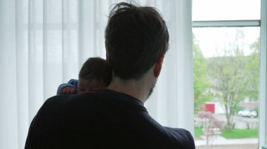 Baba, yeni doğmuş bebeği mavi bir tulumla kucağında tutuyor. Pencereden dışarı bakıyor, erken yakınlaşmanın şefkatli ve huzurlu anlarını yakalıyor ve yeni bir hayata kucak açmanın mutluluğunu yaşıyor.