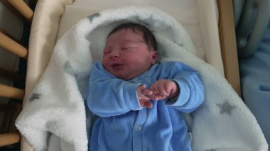 Newborn baby in blue onesie crying in crib, expression of discomfort, soft blanket, early days of life, capturing raw emotions, detailed close-up of baby's face, first moments, infancy