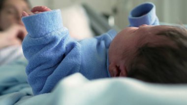 Newborn baby in a blue onesie lying on a bed in the foreground, with the mother blurred in the background in a hospital setting, highlighting the tender early moments of bonding and care