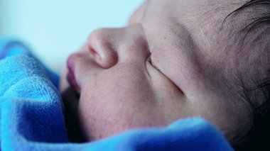 Mavi battaniyeye sarılı uyuyan yeni doğan bebeğe yakın çekim. Huzurlu yüz hatlarına ve huzurlu istirahata odaklan. Masumiyet, sükunet ve yeni hayatın güzelliği teması için ideal.