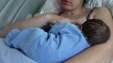 Yeni doğan bebek mavi bir battaniyeye sarılmış, anne ve çocuk arasında paylaşılan huzurlu ve besleyici anın altını çizerek, bir hastane yatağında dinlenirken annesinin kollarında emziriyor.