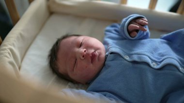 Adorable newborn in a blue onesie, lying in a crib, peacefully gazing around. Perfect for illustrating themes of infancy, parenthood, and the cozy, nurturing environment of a nursery
