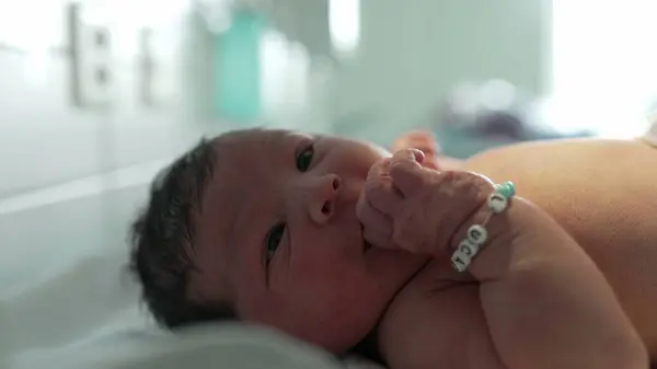 stock image Adorable newborn baby with black hair and personalized bracelet lying on hospital bed, sucking thumb, gazing curiously, peaceful expression, first moments of life, close-up