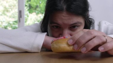 Masaya uzanmış, gözleri renkli bir çöreğe sabitlenmiş, düşünceli ve düşünceli bir ifadeyle gecikmiş haz ve cazibe temasını ileten bir kadın.