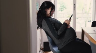 Hamile bir kadın pencerenin kenarındaki sandalyeye yaslanmış telefonuna bakıyor. Samimi ve özgün bir şekilde evinde huzurlu bir an yaşıyor.
