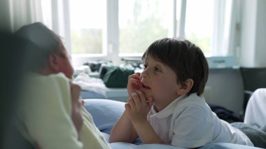 Çocuk, annesinin kollarında, hastane odasının parlak ışığıyla çevrili yeni doğmuş bebeğe merakla bakıyor. Hassas an, çocuğun büyüsünü ve ailenin yeni bebeği karşılama sevincini yansıtır.