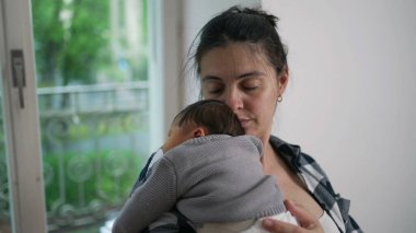 Anne, yeni doğmuş bebeğini omzunda tutuyor. Huzurlu bir bağ anında gözleri kapalı. Bebek rahat örülü bir kazak giyip huzurlu ve samimi bir atmosfer yaratıyor.