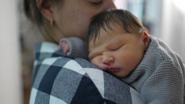 Anne, yeni doğmuş bebeğini omzunda tutarken nazikçe alnından öpüyor. Bebek rahat bir şekilde uyur, örülmüş bir süveter giyer, hassas bağlarını gösterir.