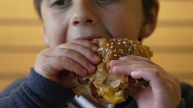 Çocuk şevkle sulu bir çizburgerden büyük bir ısırık alıyor, keyifli ve neşeli bir yüz ifadesi sergiliyor ve sıradan bir lokantada yemeğinin tadını çıkarıyor.