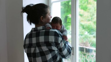 Yeni doğan bebek annesinin omzunda, ikisi de pencereden dışarı bakıyor, barışçıl bir bağ kurma anı yakalıyor. 