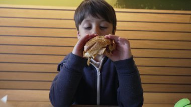 Genç çocuk büyük bir hevesle çizburgeri ısırıyor, elleri susamlı ekmeği tutuyor. İfade zevk ve açlık gösteriyor, lezzetli bir yemek yemenin basit zevkini vurguluyor.