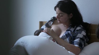 Anne bebeğini huzurlu bir yatak odasında emziriyor. Ekose gömlekli bir anne, yeni doğmuş bebeğini beyaz bir battaniyeye sarmış, huzurlu bir annelik anı yakalıyor ve onu besliyor.