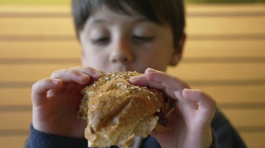 Çocuk, heyecanına ve yemeğin dokusuna odaklanmış lezzetli bir çizburgerin tadını çıkarıyor. Çocukluk sevincini ve lezzetli bir yemek yemenin basit zevklerini yakalamak için mükemmel bir an.