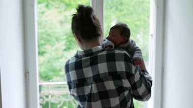 Anne, yeni doğmuş bebeğini omzuna almış pencerenin kenarında duruyor. Huzurlu bir bağ ve şefkat anı yakalıyor.