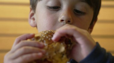 Çocuk şevkle sulu bir çizburgerden büyük bir ısırık alıyor, keyifli ve neşeli bir yüz ifadesi sergiliyor ve sıradan bir lokantada yemeğinin tadını çıkarıyor.